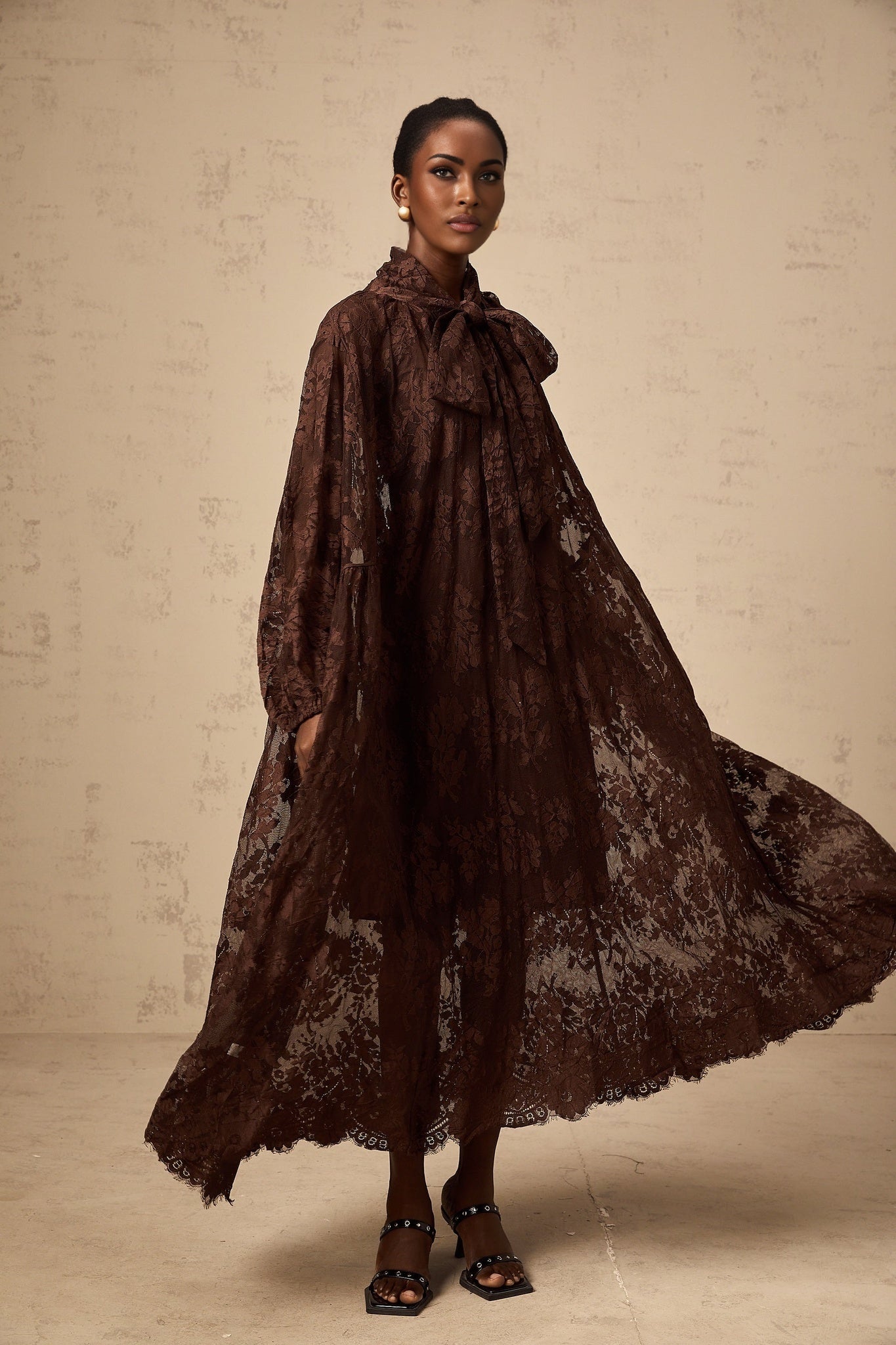 A woman wearing a brown midi dress with a flared skirt made of delicate chantilly lace The dress has a fitted bodice and falls to mid calf length The model stands in a relaxed pose showing off the elegant design of the Sylvie dress