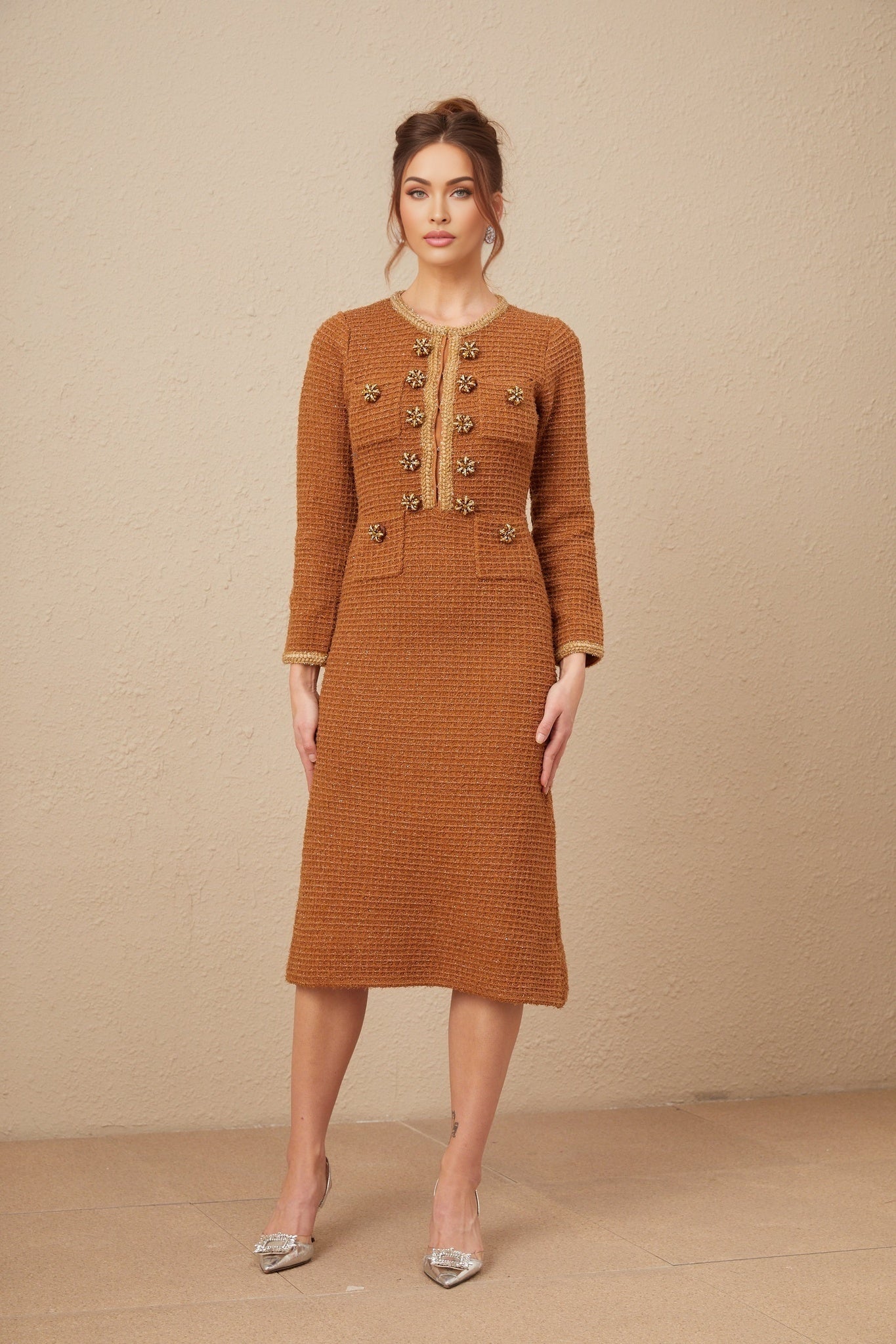 A woman wearing a brown tweed dress with braided edging standing against a plain background