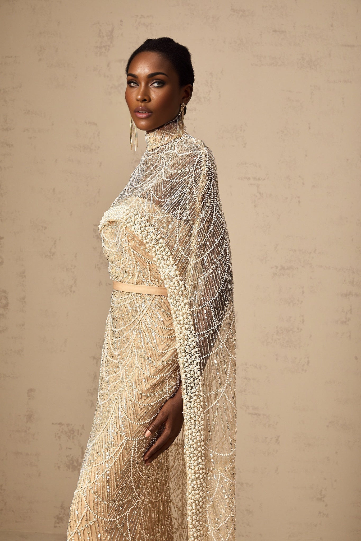 A woman wearing a champagne colored gown with a matching beaded cape The dress is floor length with a fitted silhouette while the cape drapes elegantly over the shoulders Both pieces feature intricate beadwork that sparkles in the light