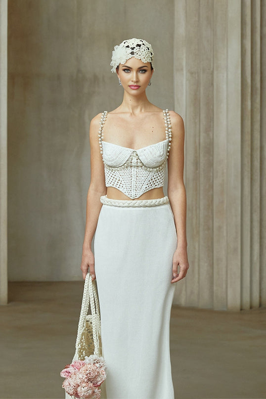 A woman wearing a cream colored crochet knit two piece outfit consisting of a short sleeve top and mini skirt standing against a plain background