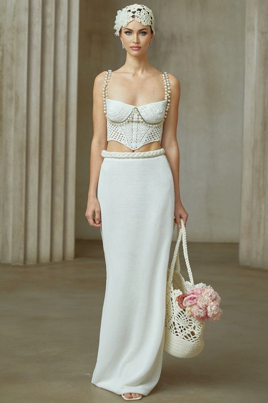 A woman wearing a cream colored crochet knit two piece set consisting of a cropped top with short sleeves and a high waisted midi skirt with a side slit