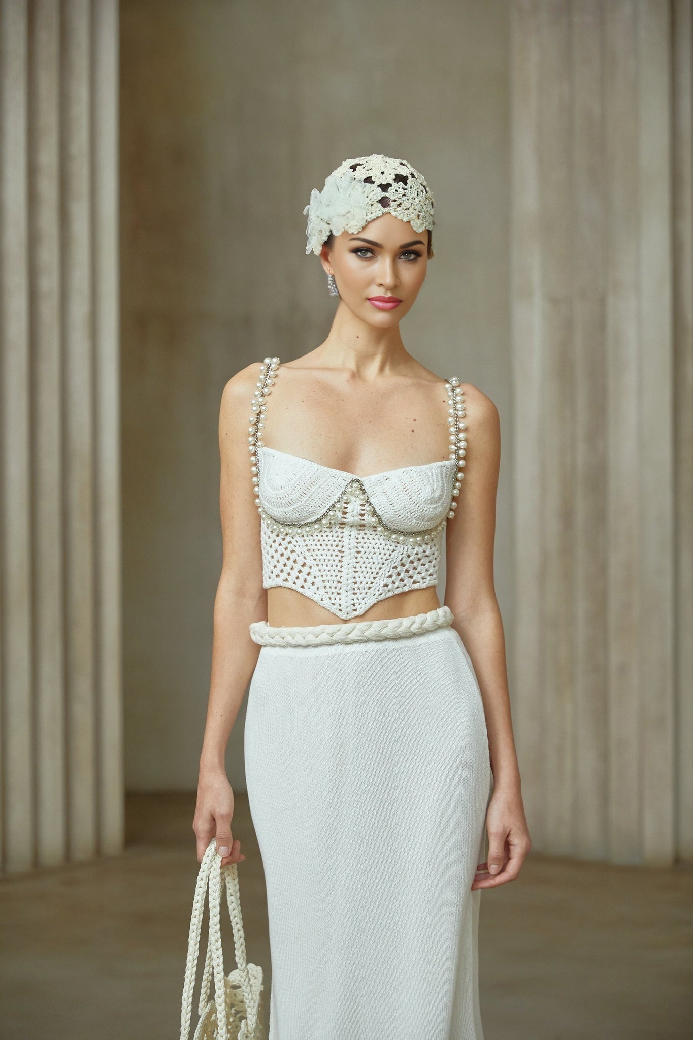 A woman wearing a crochet knit two piece set consisting of a sleeveless crop top and high waisted mini skirt in a light color standing against a plain background
