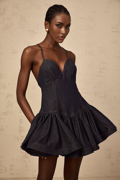 A woman wearing a dark blue denim mini dress with a flounce detail standing against a plain background