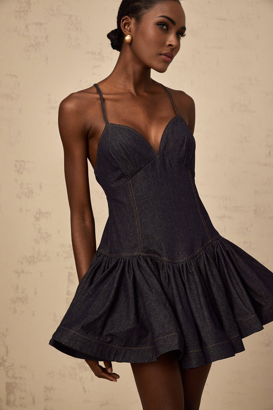 A woman wearing a dark blue denim mini dress with flounce details standing against a plain background