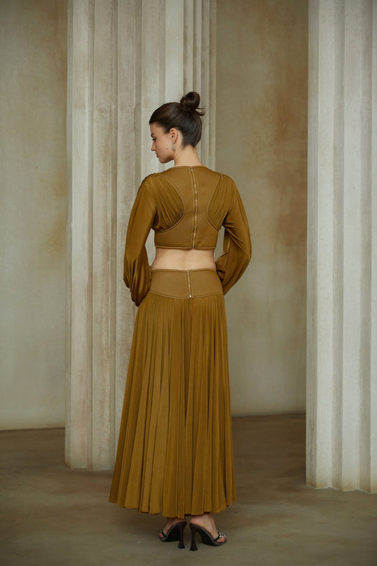 A woman wearing a green two piece outfit consisting of a fully pleated blouse and matching skirt standing against a plain background