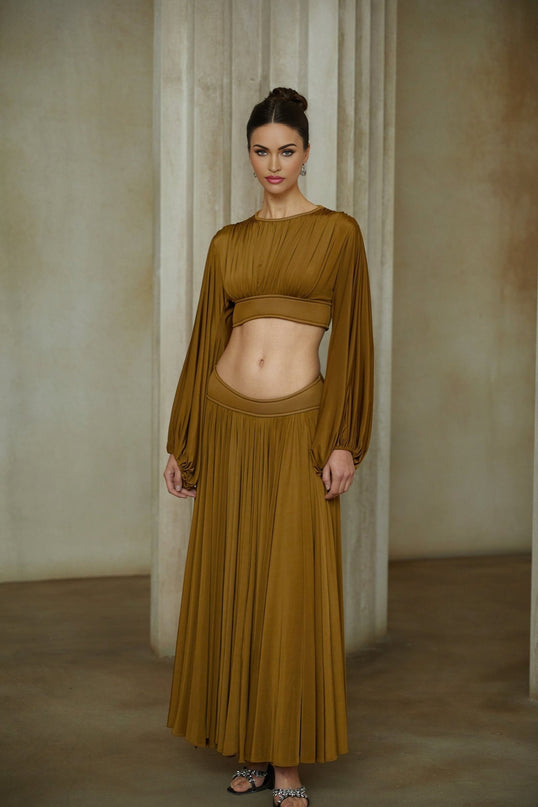 A woman wearing a green two piece outfit consisting of a pleated blouse and matching pleated skirt standing against a plain background