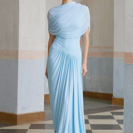 A woman wearing a light blue asymmetric pleated maxi dress with a flowing skirt and one shoulder design