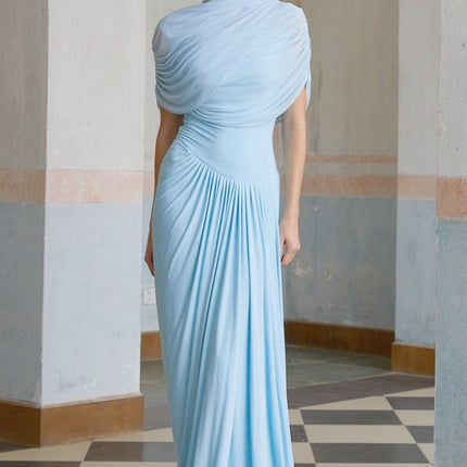 A woman wearing a light blue asymmetric pleated maxi dress with a flowing skirt and one shoulder strap