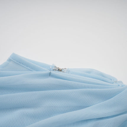 A woman wearing a light blue asymmetric pleated maxi dress with a flowing skirt and one shoulder strap