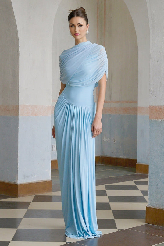 A woman wearing a light blue asymmetric pleated maxi dress with a flowy skirt and one shoulder design