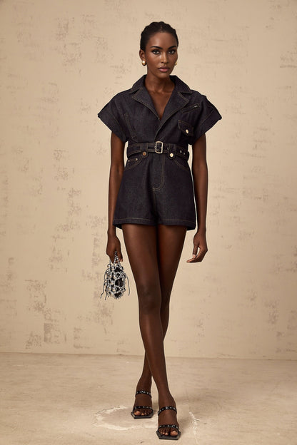 A woman wearing a light blue denim playsuit with a belted waist and short sleeves standing in a relaxed pose
