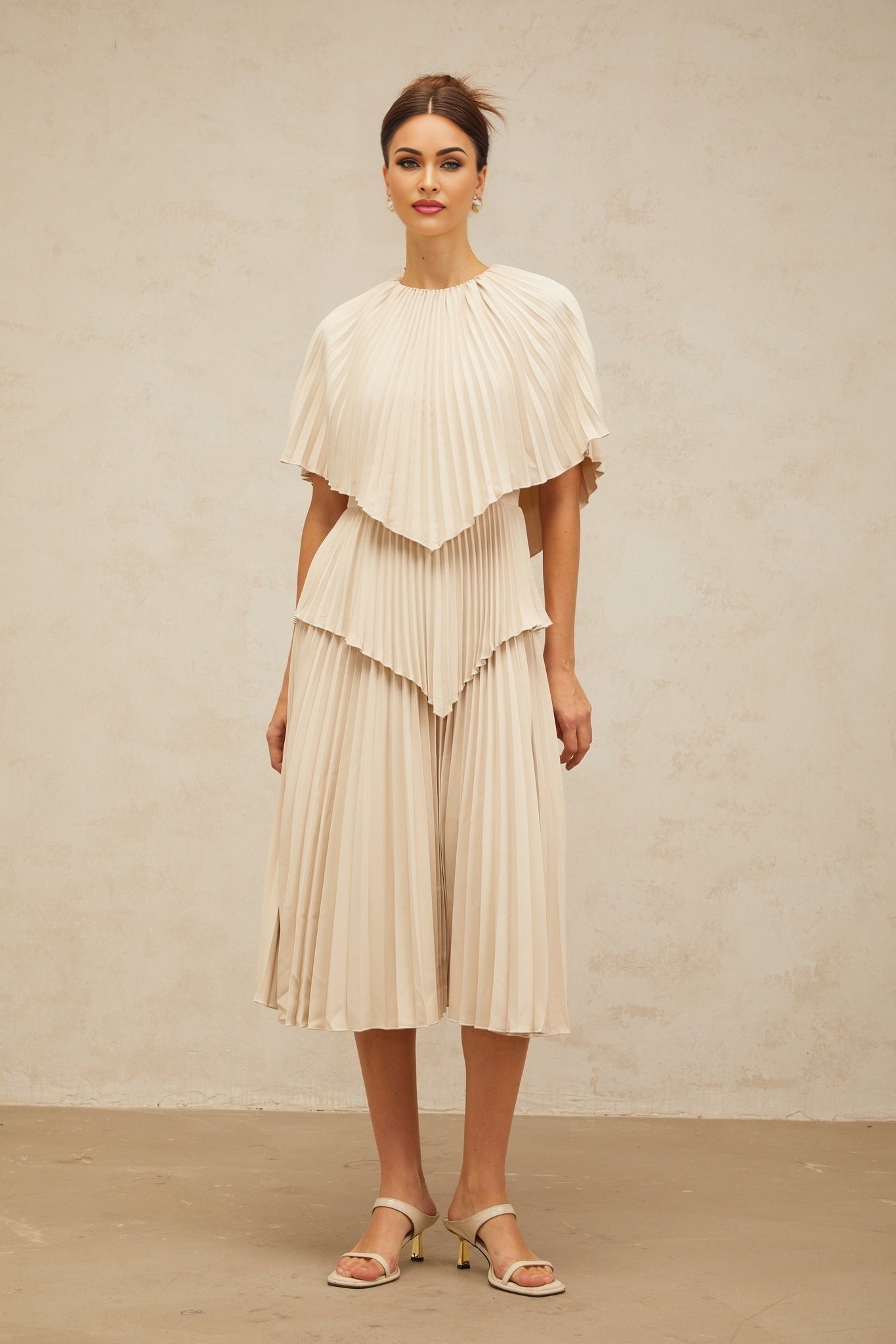 A woman wearing a light colored cape style pleated midi dress with neutral tones standing in a relaxed pose