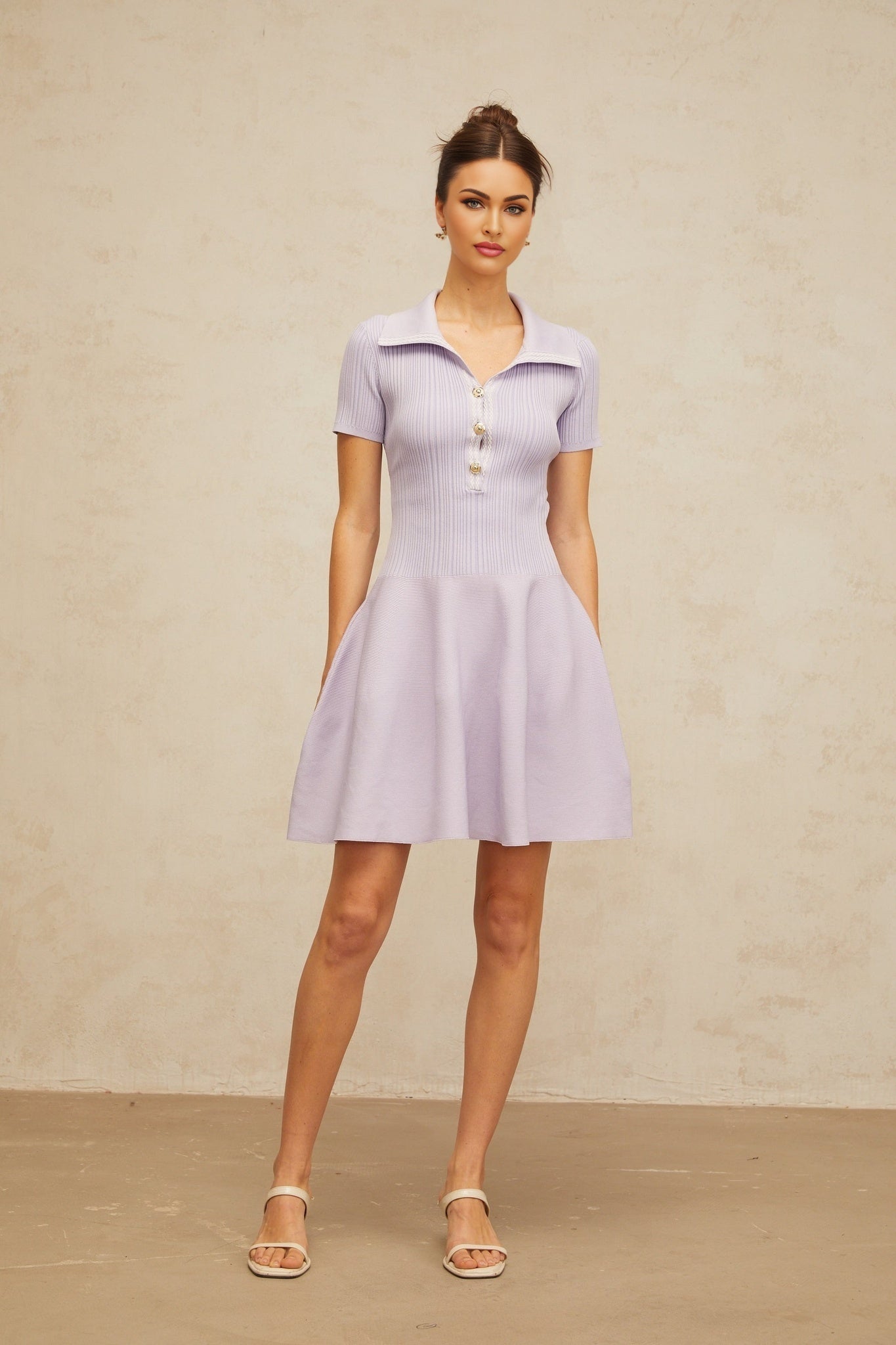 A woman wearing a light purple mini dress with a fitted bodice and flared skirt The dress features shirred detailing and is made from a knit fabric The model stands in a relaxed pose showing off the dresses flattering silhouette