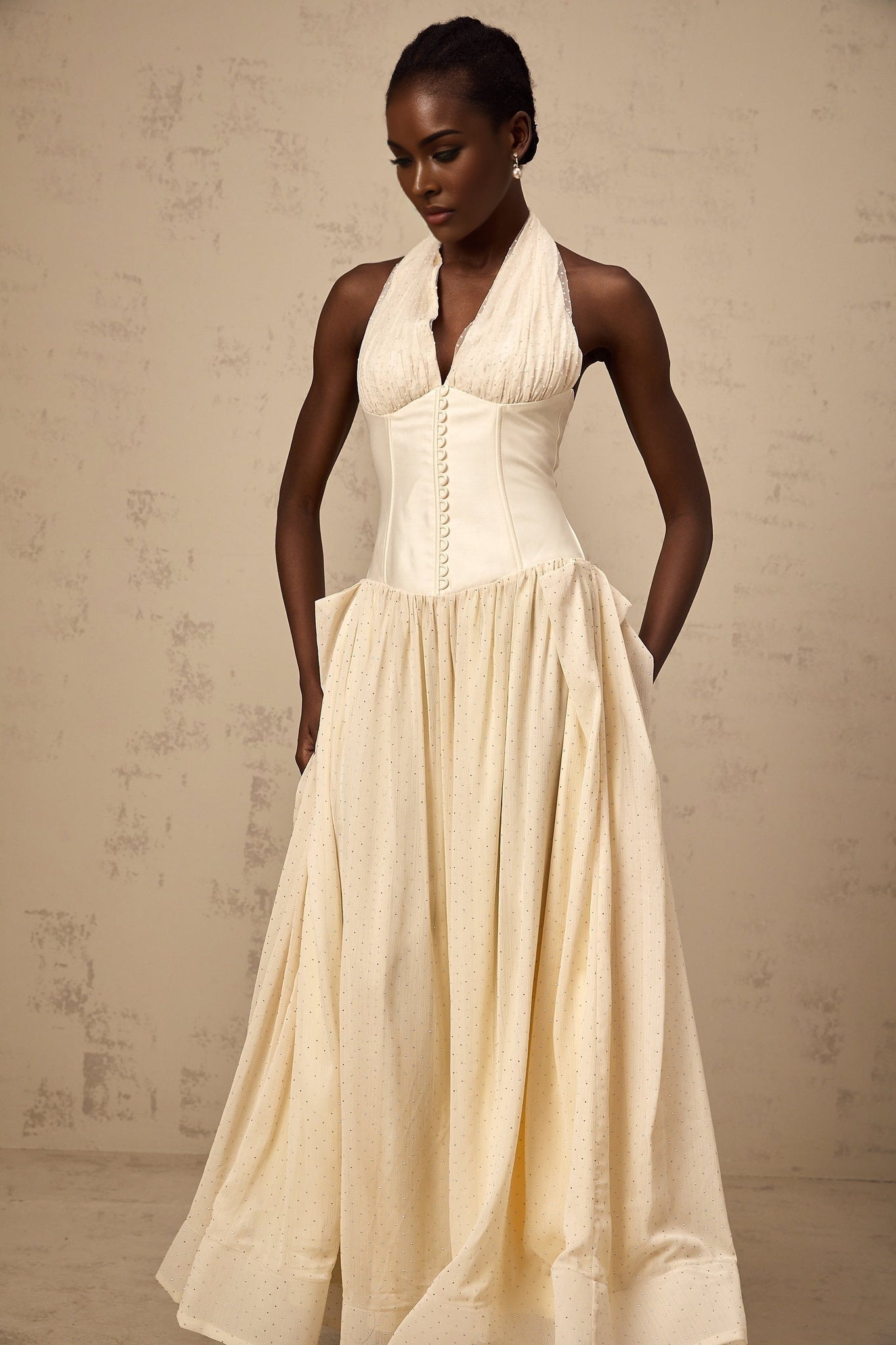 A woman wearing a lightcolored halterneck tulle midi dress with a fitted bodice and flowy skirt standing against a plain background