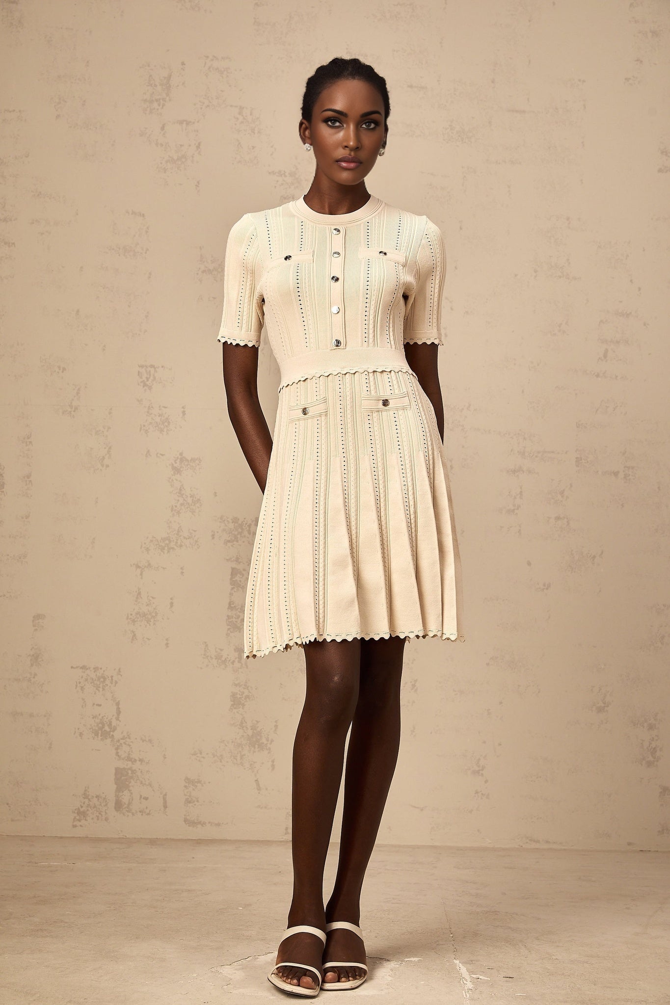 A woman wearing a lightcolored short knit dress with pleated skirt and delicate pointelle trim standing in a neutral background