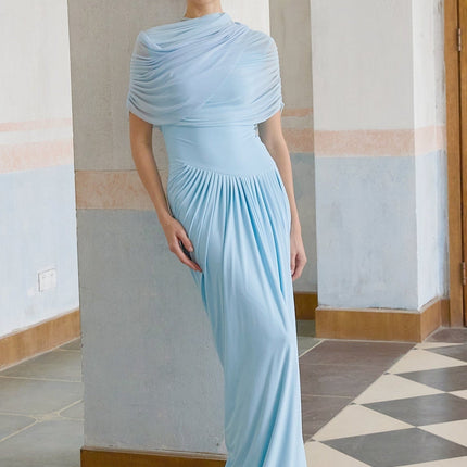 A woman wearing a long baby blue asymmetric pleated maxi dress with one shoulder strap standing against a plain background