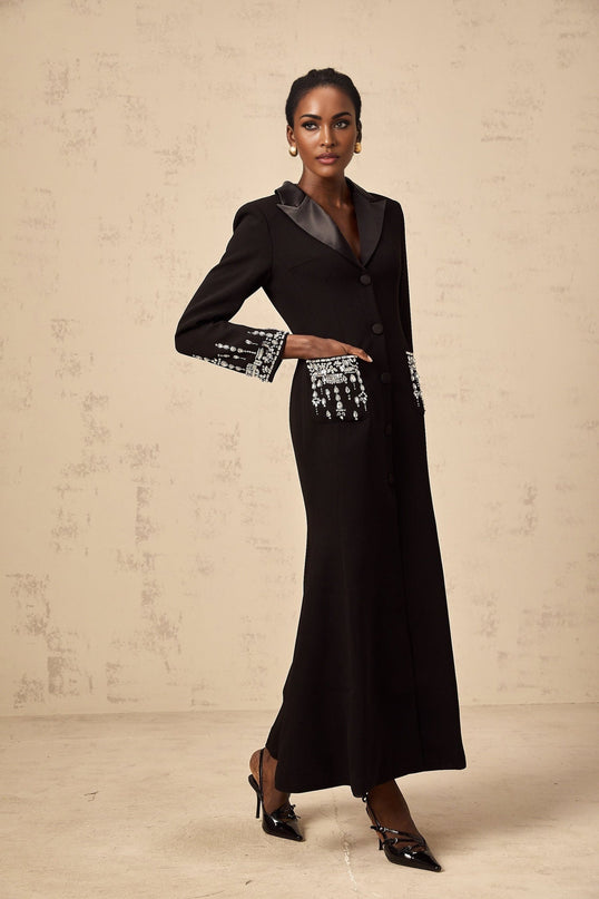 A woman wearing a long black blazer dress with embellishments and a maxi length silhouette