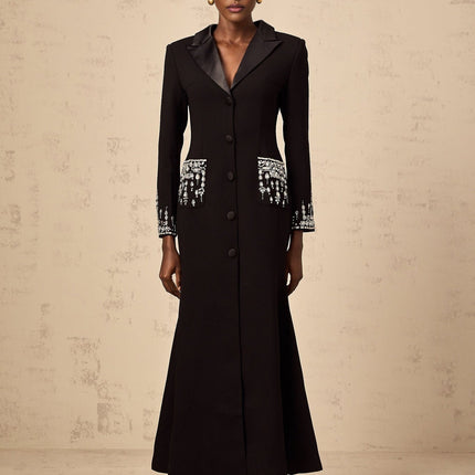 A woman wearing a long black blazer dress with embellishments standing against a plain background