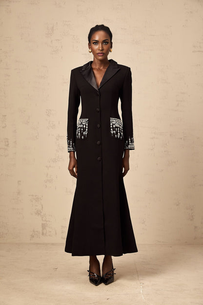 A woman wearing a long black blazer dress with embellishments standing against a plain background