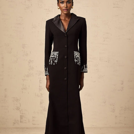 A woman wearing a long black blazer dress with embellishments standing in a neutral pose