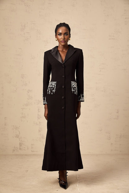 A woman wearing a long black blazer dress with embellishments standing in a neutral pose