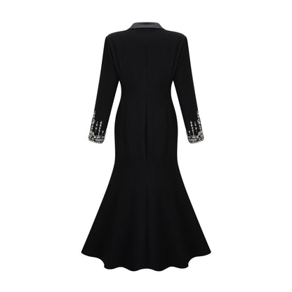 A woman wearing a long black blazer dress with embellishments standing in a relaxed pose against a plain background