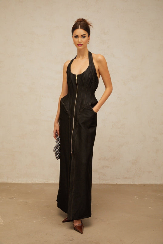 A woman wearing a long black denim maxi dress with a front slit and visible contrast stitching standing against a plain background