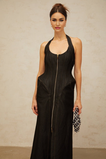 A woman wearing a long black denim maxi dress with a front slit and visible contrast stitching standing against a plain background