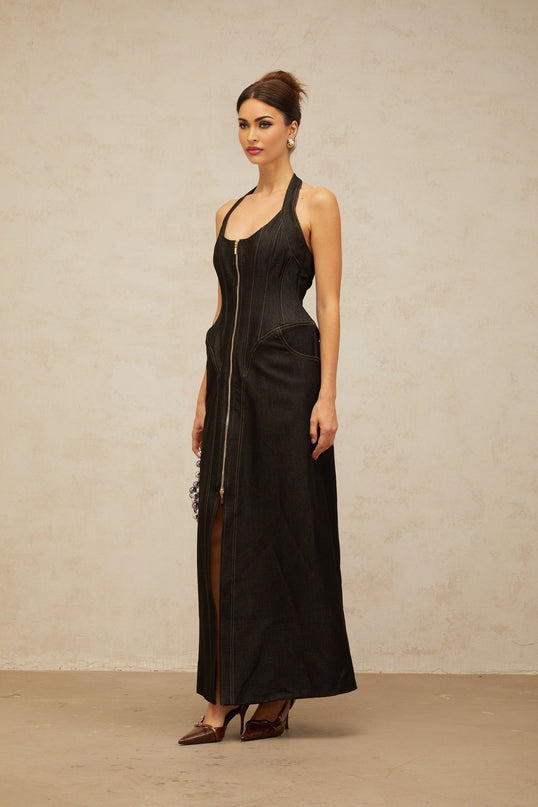 A woman wearing a long black denim maxi dress with a front slit and visible contrast stitching standing against a plain background