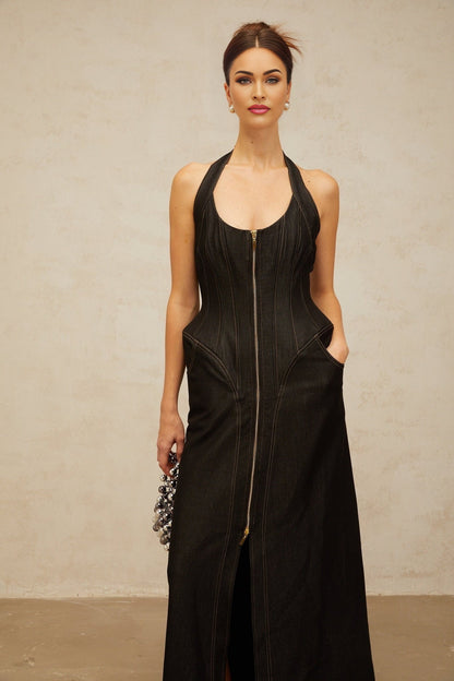 A woman wearing a long black denim maxi dress with a front slit and visible contrast stitching standing in a casual pose