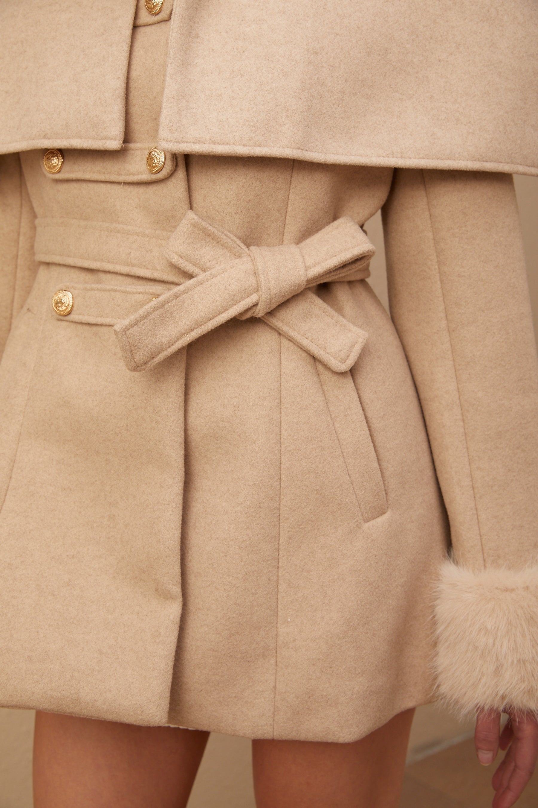 A woman wearing a long black faux fur coat with a detachable cape collar standing against a plain background