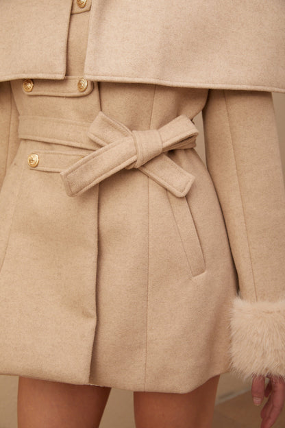 A woman wearing a long black faux fur coat with a detachable cape collar standing against a plain background