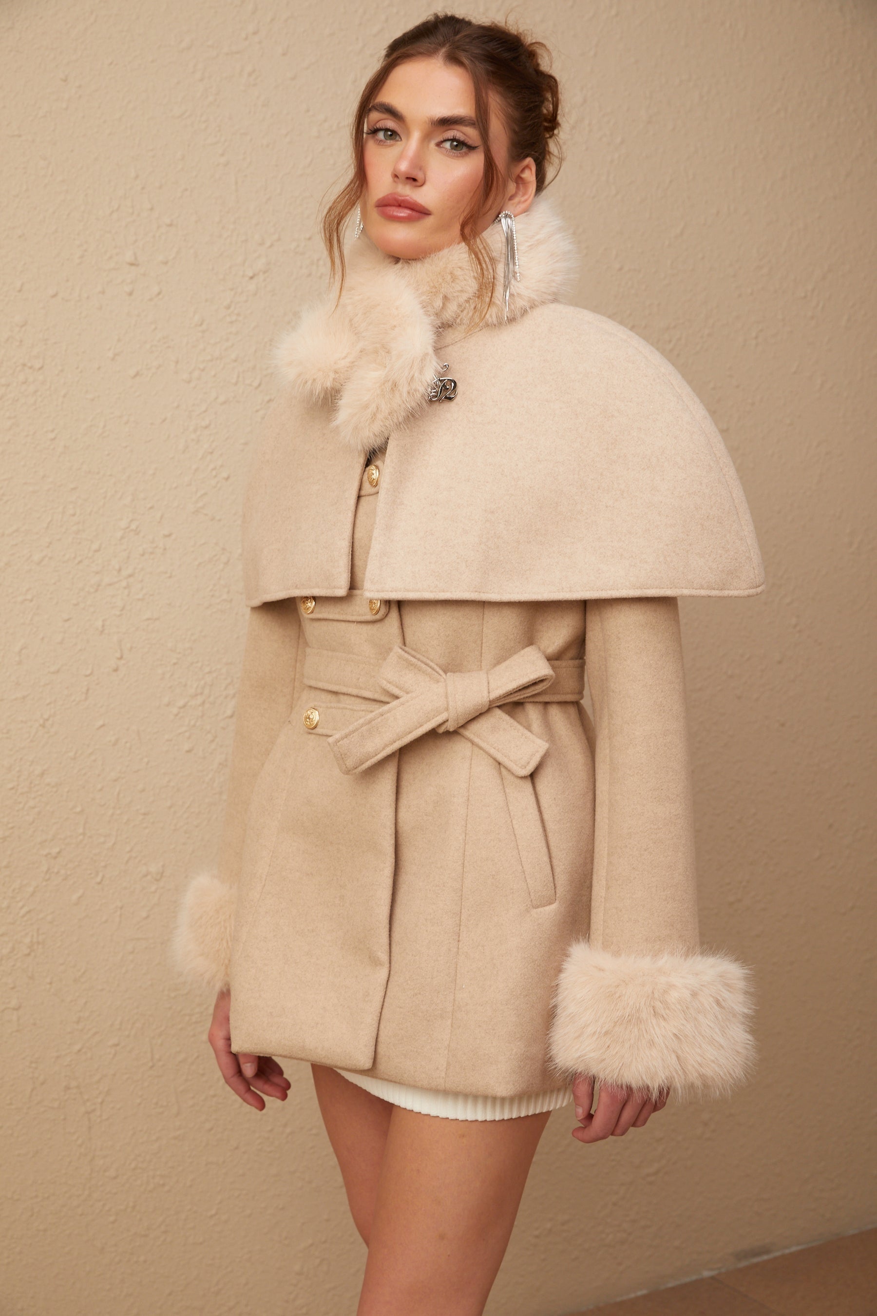 A woman wearing a long black faux fur coat with a detachable cape draped over her shoulders standing against a plain background
