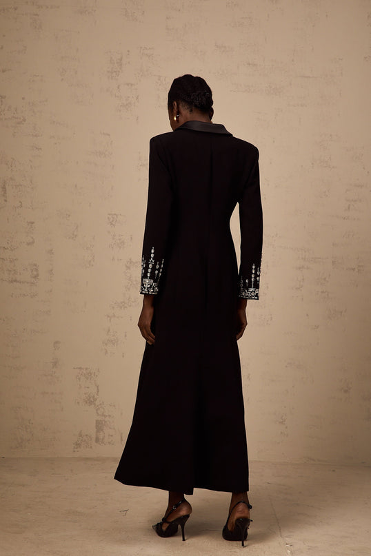A woman wearing a long black maxi blazer dress with embellished details standing in a neutral pose