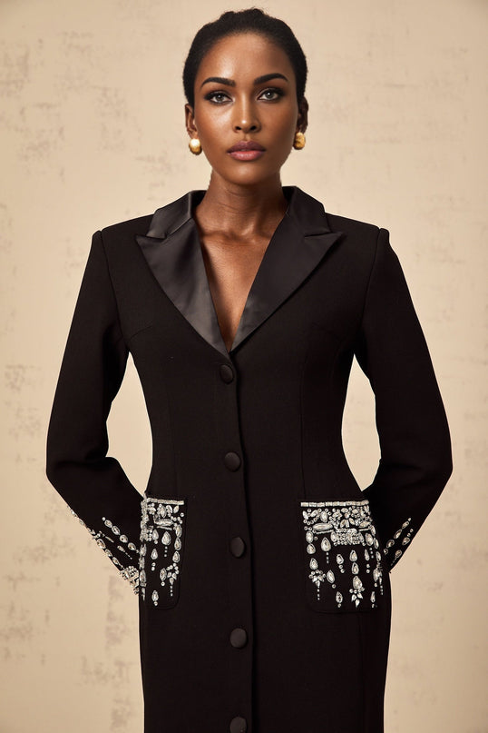 A woman wearing a long black maxi blazer dress with embellishments standing in a neutral setting