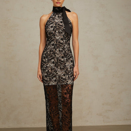 A woman wearing a long black maxi dress with floral lace pattern and halter neckline standing against a plain background