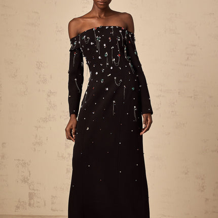 A woman wearing a long black offshoulder maxi dress adorned with crystal embellishments posing elegantly against a plain background
