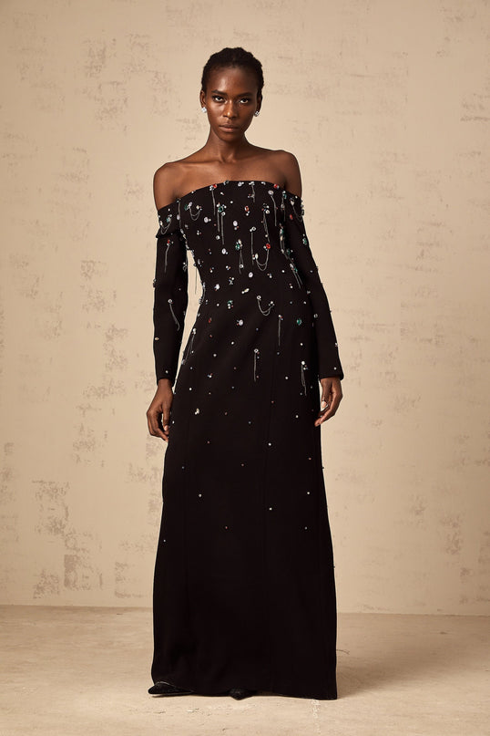 A woman wearing a long black offshoulder maxi dress adorned with crystal embellishments posing elegantly against a plain background