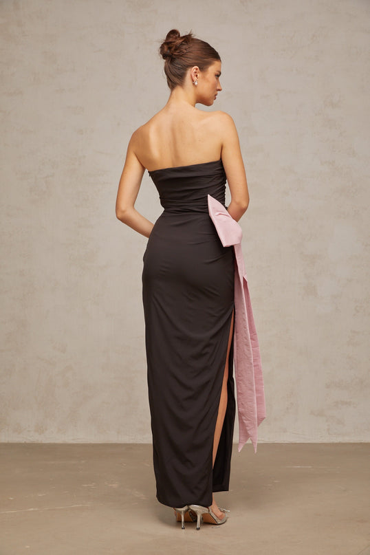 A woman wearing a long black offshoulder maxi dress with a bow detail at the neckline