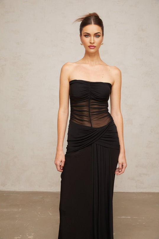A woman wearing a long black semi sheer maxi dress with pleated tulle fabric standing in a neutral pose
