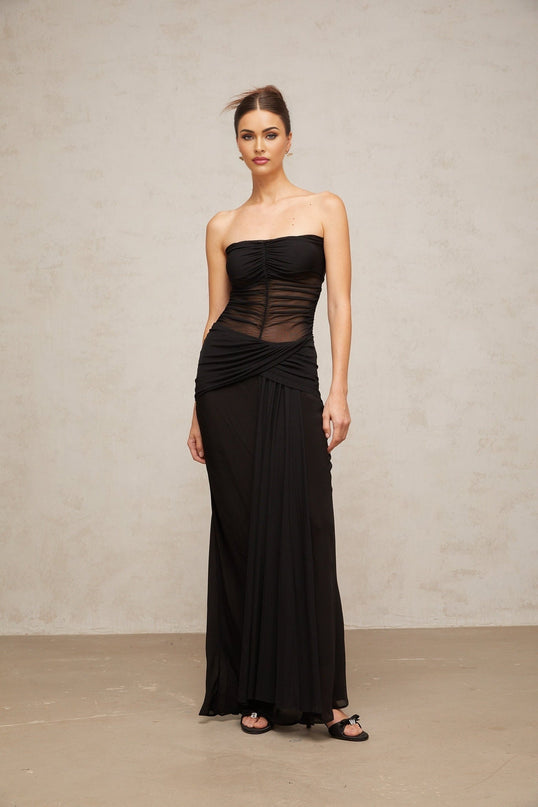 A woman wearing a long black semi sheer pleated tulle maxi dress with a fitted bodice and flowing skirt