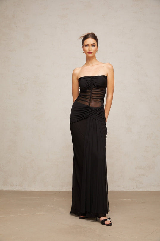 A woman wearing a long black semi sheer pleated tulle maxi dress with a fitted bodice and flowing skirt