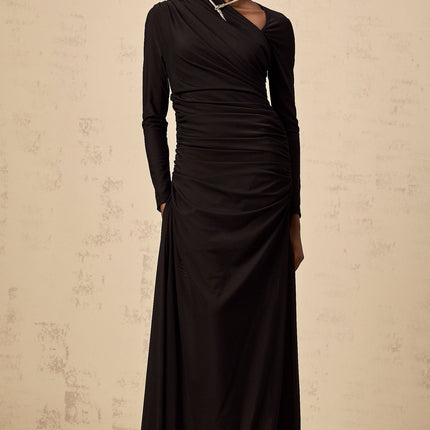 A woman wearing a long black tulle dress with an asymmetric hemline and cutout details standing against a plain background