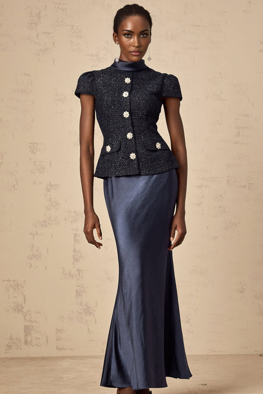 A woman wearing a long blue dress with a fitted bodice and peplum waist The dress features panels and extends to floor length The model has her hair down and is posing in front of a plain background