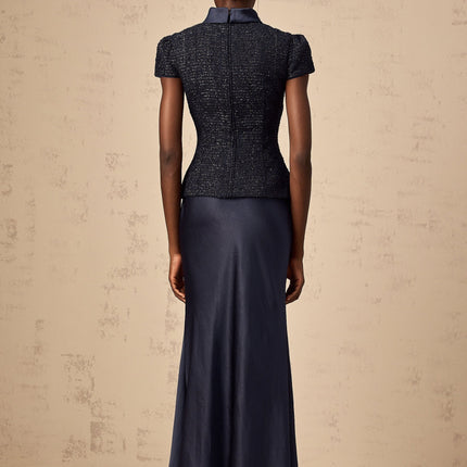 A woman wearing a long blue dress with a fitted waist and flared bottom standing against a plain background The dress features a peplum detail at the waist and panelled construction throughout its length