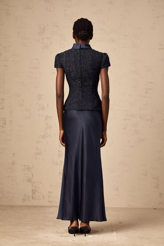 A woman wearing a long blue dress with a fitted waist and flared bottom standing against a plain background The dress features a peplum detail at the waist and panelled construction throughout its length