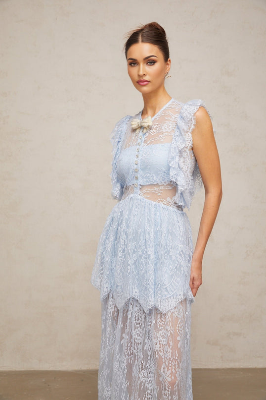 A woman wearing a long blue lace maxi dress with layered skirt and delicate floral pattern standing against a plain background