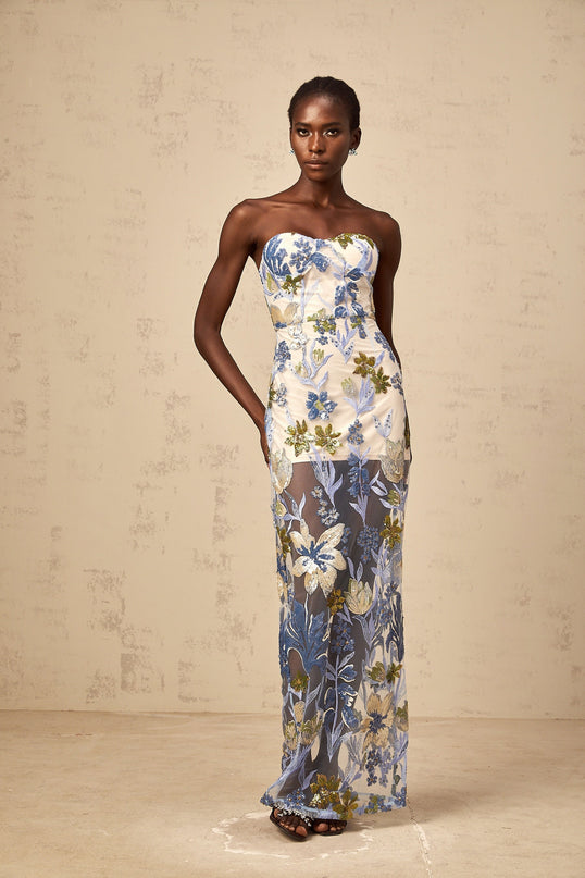 A woman wearing a long blue maxi dress with a corset bodice featuring sequined flower embroidery and a flowing tulle skirt