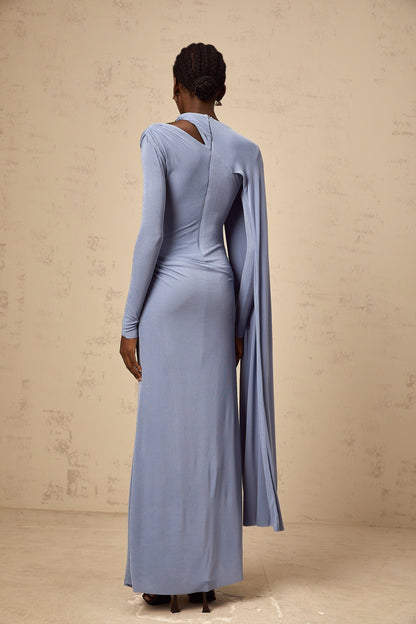 A woman wearing a long blue maxi dress with a fitted silhouette and a diagonal shawl detail across the bodice