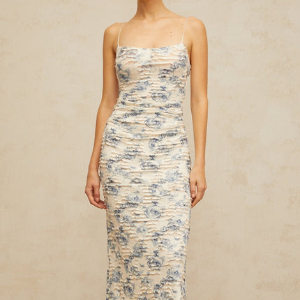 A woman wearing a long blue maxi dress with a textured floral print pattern standing against a plain background